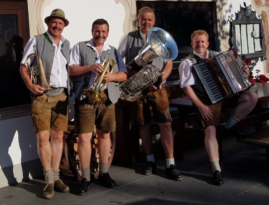 Vier Männer in traditionellen bayerischen Trachten stehen lächelnd zusammen. Sie halten Musikinstrumente wie eine Tuba, ein Akkordeon und Trompeten.