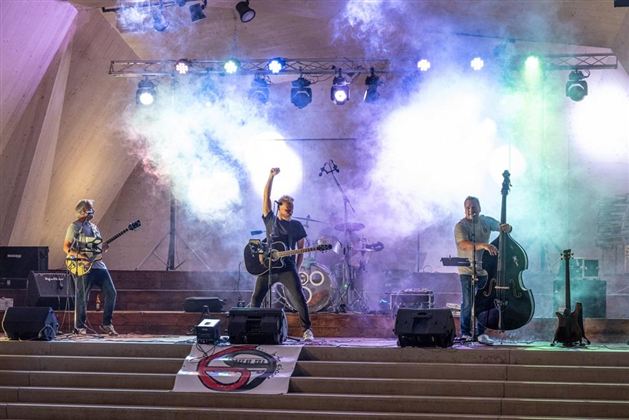 Eine Band spielt auf einer Bühne mit bunten Lichtern und Nebel. Der Sänger hebt die Hand, während die Musiker mit ihren Instrumenten auftreten.