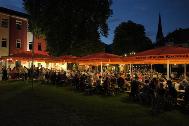 A lively outdoor event with many people sitting at tables under orange umbrellas. It is night, and the surroundings are softly illuminated.