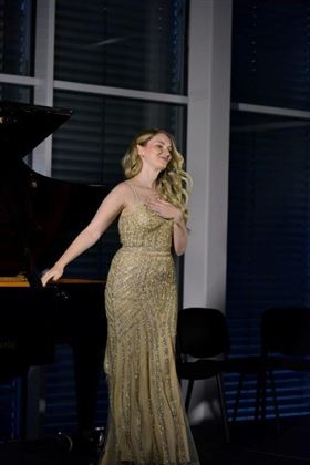 En sangerinde i en glitrende guldkjole står på scenen. I baggrunden ses et klaver og store vinduer.