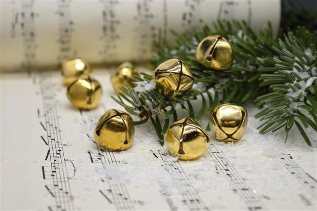 Eine Vielzahl von goldenen Glocken liegt auf einem Blatt mit Noten. Dabei sind auch Tannenzweige und etwas Schnee zu sehen.