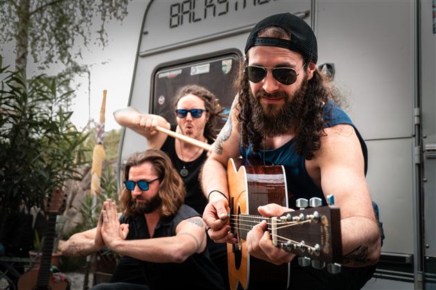Drie muzikanten spelen in een ontspannen sfeer. Op de achtergrond zijn planten en een caravan te zien.