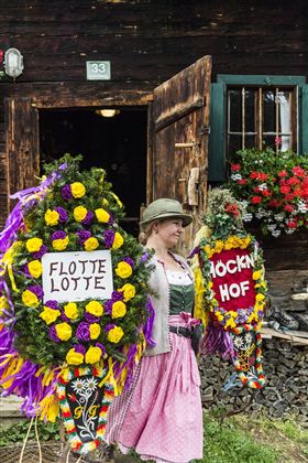 Een vrouw in traditionele klederdracht staat voor een houten huis en draagt twee feestelijk versierde kransen. De kransen zijn met kleurrijke bloemen versierd en dragen de teksten "FLOTTEN LOTTE" en "HÖCKEN HOF".