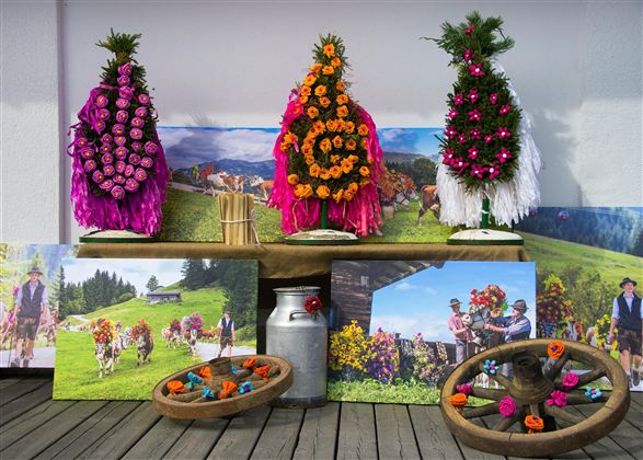 Traditionelle Blumenkränze in bunten Farben stehen auf einem Tisch. Im Hintergrund sind Bilder von der Alm und fröhlichen Menschen mit Kühen zu sehen.