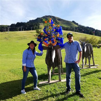 Ein Paar steht neben einer Skulptur einer Kuh, die mit bunten Blumen geschmückt ist. Im Hintergrund sind sanfte Hügel und eine klare Himmel zu sehen.