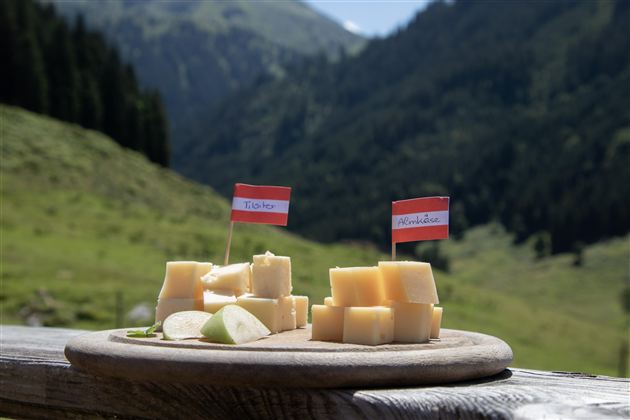 Ein elegantes Käsebrett mit verschiedenen Käsestücken, dekoriert mit kleinen Flaggen. Im Hintergrund sind grüne Berge und eine natürliche Landschaft zu sehen.