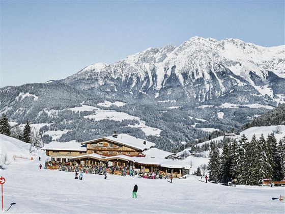 A picturesque winter landscape with snow-covered mountains. In the foreground stands a mountain cabin, surrounded by skiers and fir trees.