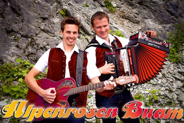 Two musicians in traditional attire stand in front of a rock wall. One plays the guitar, the other the accordion.
