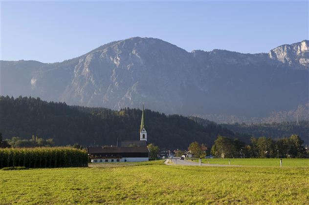 Eine malerische Landschaft mit Bergen im Hintergrund. Im Vordergrund steht eine kleine Kirche umgeben von Wiesen.