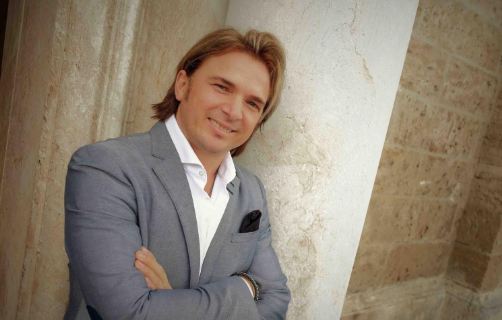 Ein lächelnder Mann steht an einer Säule mit gekreuzten Armen. Er trägt einen blauen Anzug und wirkt selbstbewusst und freundlich.