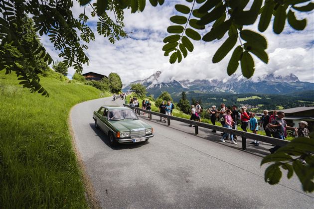 Een groep mensen wandelt over een pad door een groen landschap. Op de achtergrond zijn zachte heuvels en een klein dorp zichtbaar.
