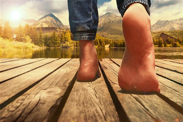 Osoba jde naboso po dřevěné lávce podél jezera. V pozadí jsou hory a slunečné nebe.