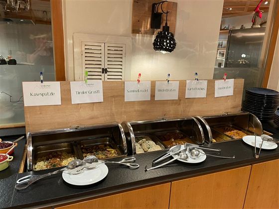 Ein Buffet mit verschiedenen Speisen in warmen Behältern. Darüber hängen Beschilderungen mit den Namen der Gerichte.