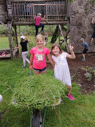 Zwei Mädchen stehen lächelnd vor einer Schubkarre mit frischem Gras. Im Hintergrund spielen andere Kinder auf einem Spielplatz.