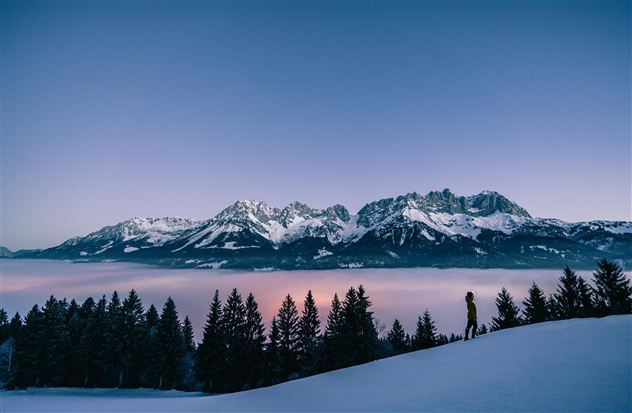 Eine beeindruckende Winterlandschaft mit schneebedeckten Bergen und einem hohen Aussichtspunkt. Der Himmel ist in sanften Farben beleuchtet und Nebel zieht durch das Tal.