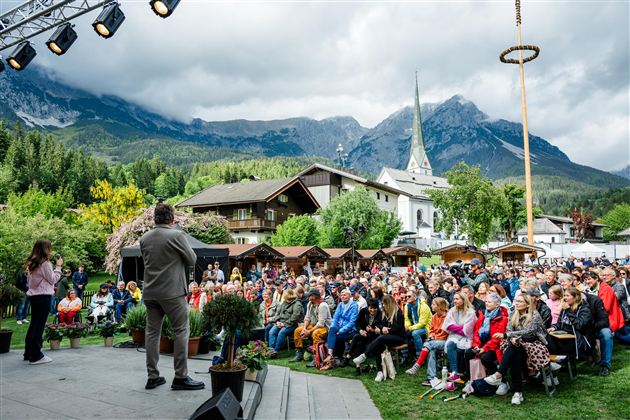 Eine große Zuschauermenge sitzt im Freien vor einer Bühne, während ein Redner spricht. Im Hintergrund sind Berge und eine Kirche zu sehen, was eine malerische Kulisse schafft.
