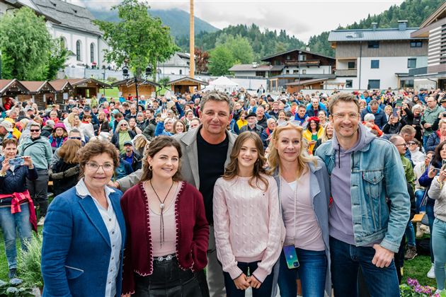 Eine Gruppe von fünf Personen steht lächelnd vor einer großen Menschenmenge. Im Hintergrund sind Berge und Gebäude zu sehen.