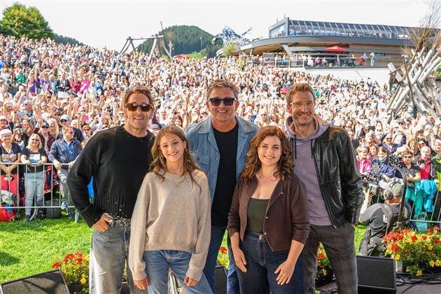 Fünf Personen stehen lächelnd vor einer großen Menschenmenge. Im Hintergrund sind Berge und ein Gebäude zu sehen.
