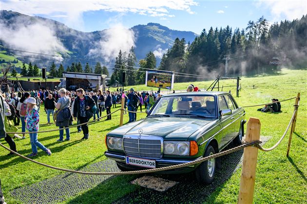 Ein klassisches Auto steht auf einem grünen Feld. Im Hintergrund sind Menschen und Berge zu sehen, und die Atmosphäre wirkt festlich.