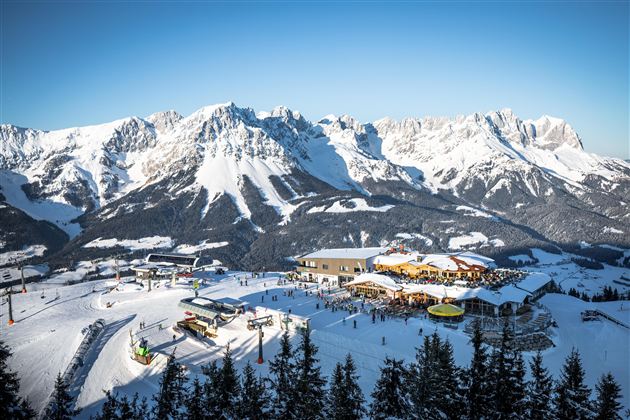 A beautiful snowy landscape with mountains in the background. Skiers and a mountain station can be seen in the foreground.