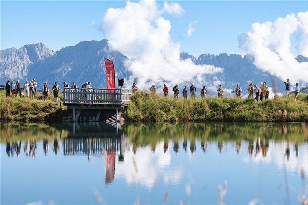 Eine Gruppe von Menschen steht auf einer Plattform am Wasser. Im Hintergrund sind majestätische Berge und ein blauer Himmel mit weißen Wolken zu sehen.