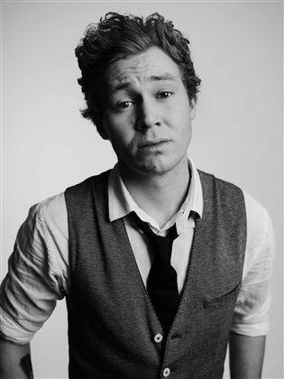 Ein junger Mann trägt eine Weste und ein Hemd. Er schaut in die Kamera mit einem neutralen Gesichtsausdruck.