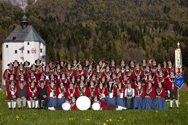 Eine große Gruppe von Menschen in traditioneller Kleidung steht im Freien vor einem historischen Gebäude. Die Teilnehmer lächeln und posieren gemeinsam auf einer Wiese mit grünen Bäumen im Hintergrund.