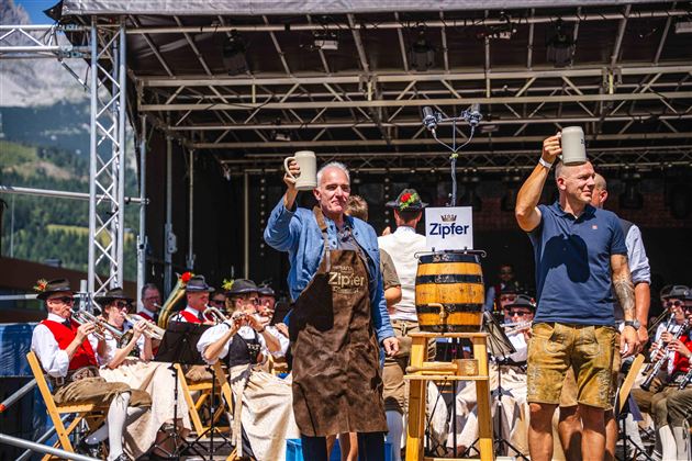 Zwei Männer heben Bierkrüge auf einer Bühne während einer Feier. Im Hintergrund sind Musiker in traditionellen Kostümen zu sehen.