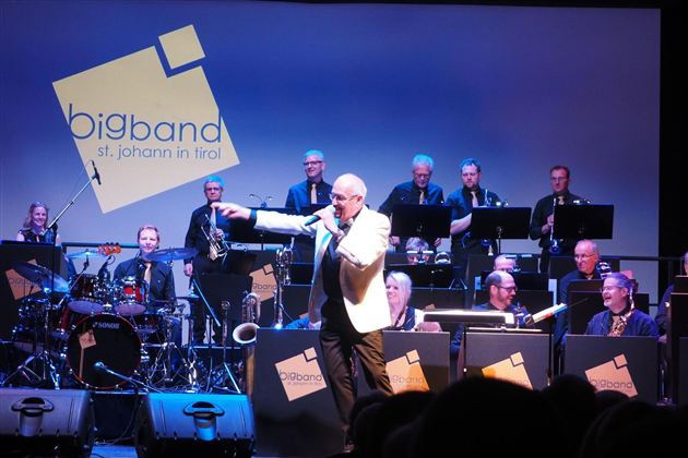 Ein Sänger tritt auf der Bühne auf, während eine Bigband hinter ihm spielt. Die Musiker sind in formellen Kleidern und konzentrieren sich auf ihre Instrumente.