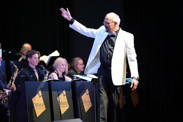 Ein Dirigent in einem weißen Smoking leitet ein Orchester. Im Hintergrund sind Musiker mit ihren Instrumenten zu sehen.