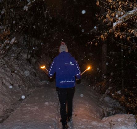 Eine Person läuft mit Fackeln auf einem verschneiten Weg in der Nacht. Es fällt Schnee und die Umgebung ist dunkel und ruhig.