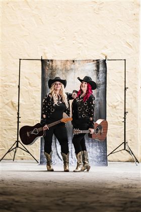 Zwei Musikerinnen stehen vor einem Hintergrund und halten Gitarren. Sie tragen schwarze Outfits und Hüte.