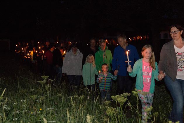 Een groep mensen loopt 's nachts, allemaal met kaarsen in de hand. Op de achtergrond zijn bomen en gras te zien.