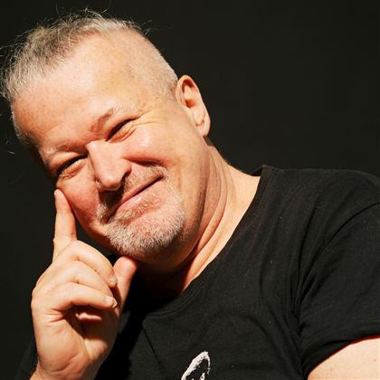 Ein lächelnder Mann mit kurzen, grauen Haaren und einem Bart. Er trägt ein schwarzes T-Shirt und hat eine nachdenkliche Pose.