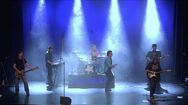 Een band speelt live op het podium met kleurrijk licht en mist. De muzikanten zijn in levendige poses en gebruiken verschillende instrumenten.