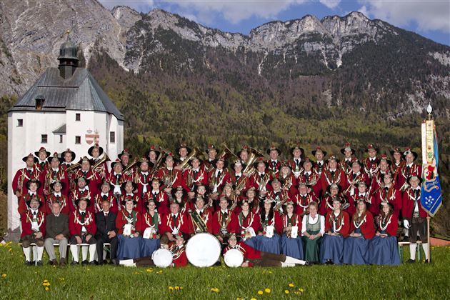 Eine große Gruppe von Menschen in traditionellen Trachten steht vor einer malerischen Berglandschaft. Im Hintergrund sieht man ein historisches Gebäude und eine Fahne.