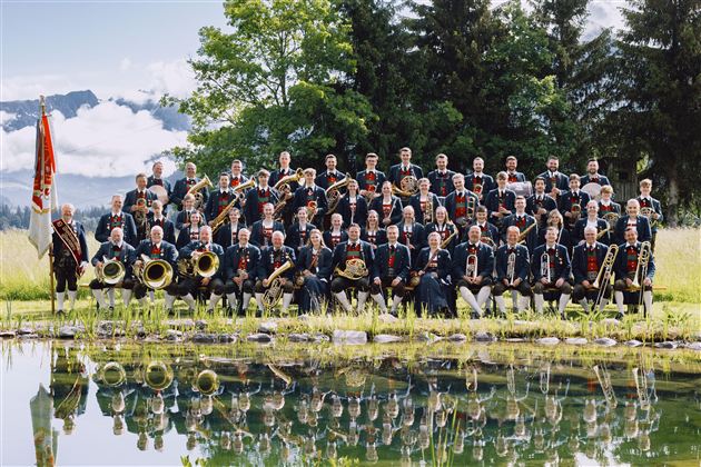 En stor brassband står foran en dam i naturen. I baggrunden kan man se bjerge og en klar himmel.