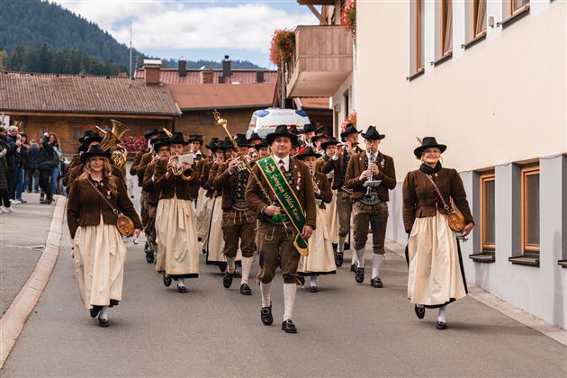A traditional costume parade with people in festive clothing is walking through a street. In the foreground, a man with a flag leads the group.