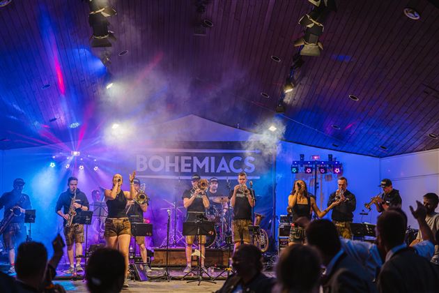 Eine lebhafte Band spielt auf einer Bühne, umgeben von bunten Lichtern und Nebeleffekten. Das Publikum ist begeistert und applaudiert.