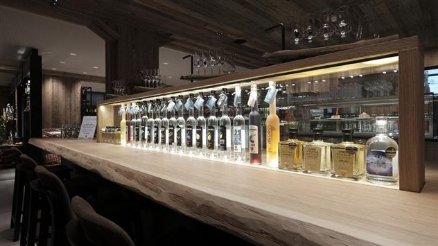 Eine stilvolle Bar mit einer langen Holztheke und beleuchtetem Regal. Mehrere Flaschen Spirituosen stehen ordentlich hinter der Theke.