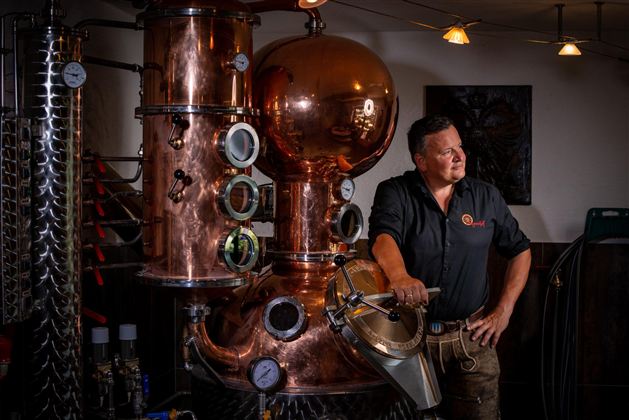 Ein Mann steht neben einer großen Kupferdestille in einer Brennerei. Die Umgebung hat eine rustikale Atmosphäre mit verschiedenen Geräten und einem industriellen Look.