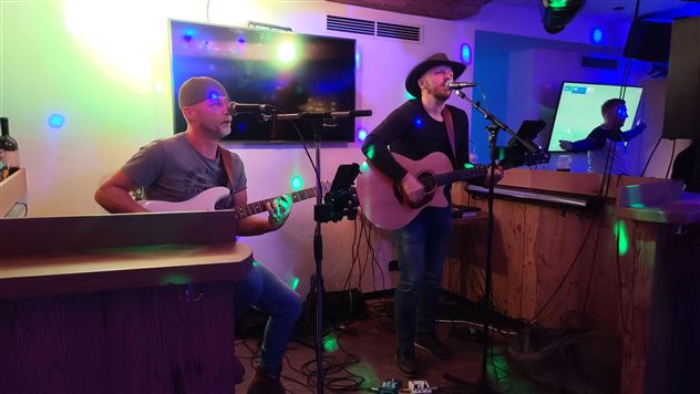 A musician plays guitar and sings live, while another musician accompanies on bass. The setting is illuminated with colorful lights.