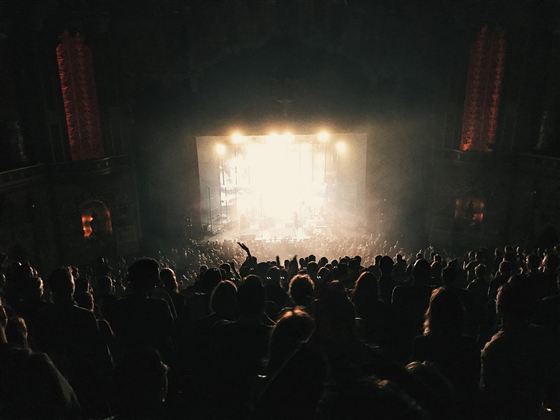 Eine große Menschenmenge bei einem Konzert in einem dunklen Saal. Helles Licht strahlt von der Bühne und schafft eine aufregende Atmosphäre.