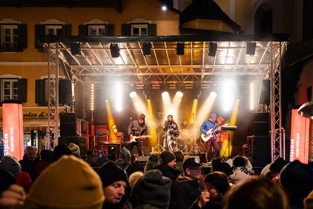 Een liveband speelt op een podium voor enthousiast publiek. De sfeer is feestelijk met lichten en een levendige achtergrond.