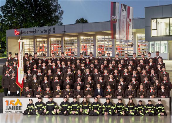 Eine große Gruppe von Feuerwehrleuten steht vor einem Feuerwehrgebäude. Sie tragen Uniformen und lächeln in die Kamera.