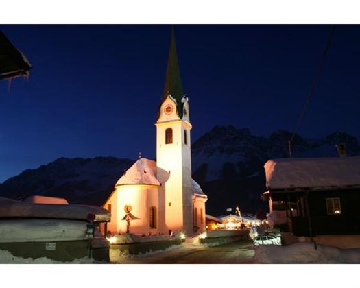 Eine charmante Kirche mit einem hohen, grünen Turm, umgeben von Schnee. Die Szenerie wird von einem klaren Nachthimmel und sanften Beleuchtungen der Gebäude geprägt.