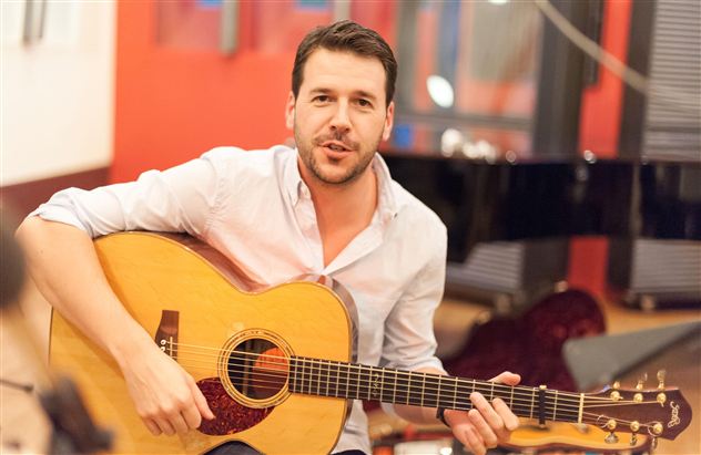 Ein Mann sitzt mit einer Gitarre in der Hand und lächelt. Im Hintergrund ist ein Klavier zu sehen.
