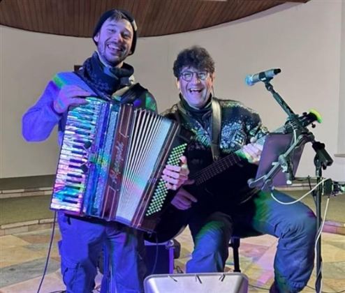 Zwei Musiker stehen in einer Innenraum-Umgebung und spielen gemeinsam Werke. Einer spielt ein Akkordeon, während der andere eine Gitarre hält und lächelt.