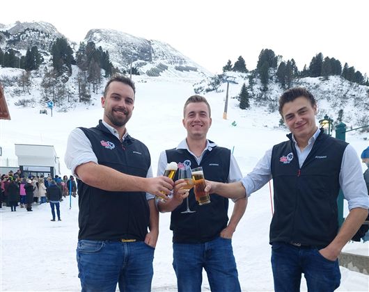 Drei Männer stoßen mit Gläsern auf einer schneebedeckten Fläche an. Im Hintergrund sind Berge und eine Menschenmenge zu sehen.