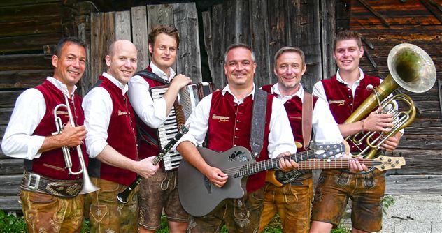 Sechs Musiker in traditionellen Trachten stehen vor einer Holzwand. Sie halten verschiedene Instrumente und lächeln in die Kamera.
